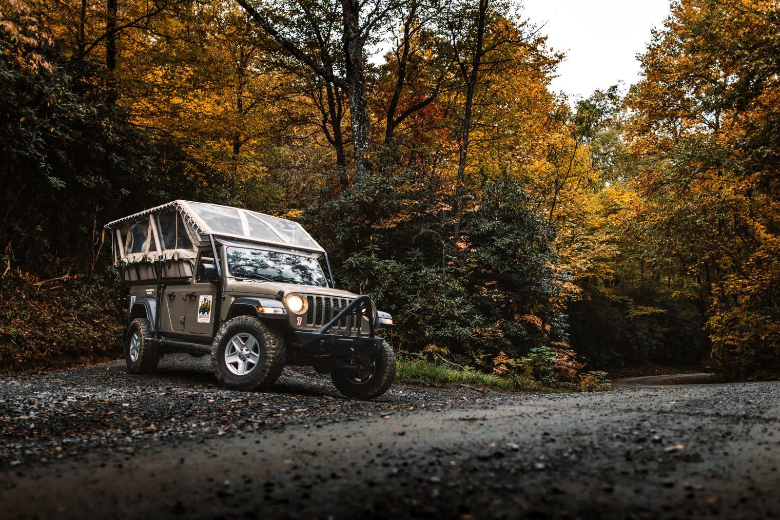 Open-air Asheville tour Jeep parked on a wet gravel forest road in Pisgah National Forest under gold and orange autumn hardwoods