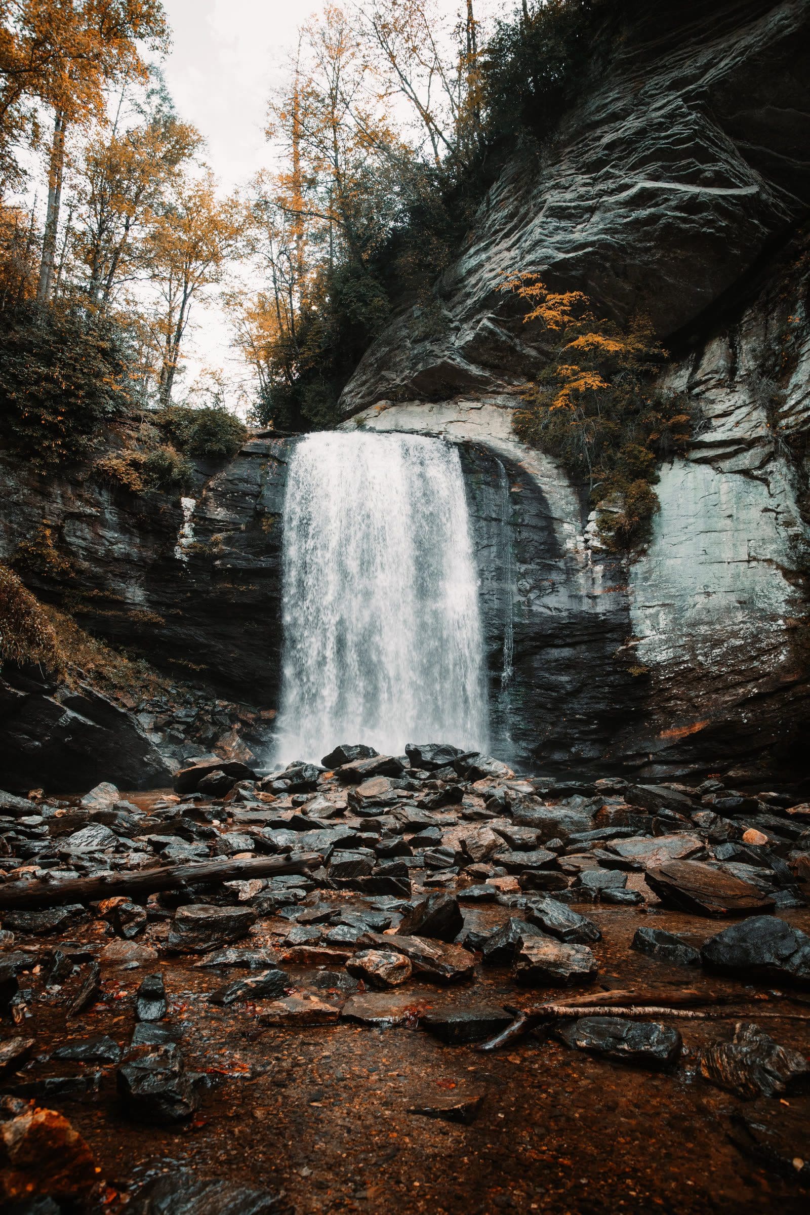 Looking Glass Falls photographed from creek level in Pisgah National Forest, the falls dropping into a shallow pool across a field of wet angular slate boulders in late autumn