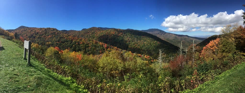 A wide view from a Blue Ridge Parkway overlook showing a valley of mixed autumn color, with orange and rust patches among still-green forest running up to a large ridge under a deep blue sky