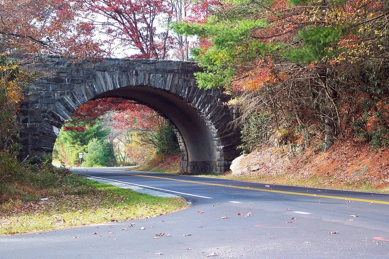 A stone arch overpass on the Blue Ridge Parkway in autumn, the two-lane road curving beneath it through red and gold foliage